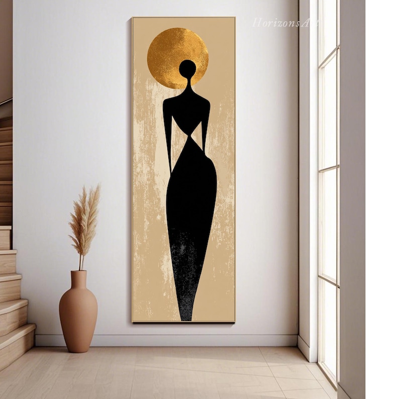 Puede incluir: Impresi&oacute;n art&iacute;stica abstracta que presenta una silueta negra de una figura con un halo dorado sobre un fondo beige. La figura lleva un vestido largo negro. La obra de arte est&aacute; enmarcada en un borde dorado y se exhibe en una habitaci&oacute;n bien iluminada.