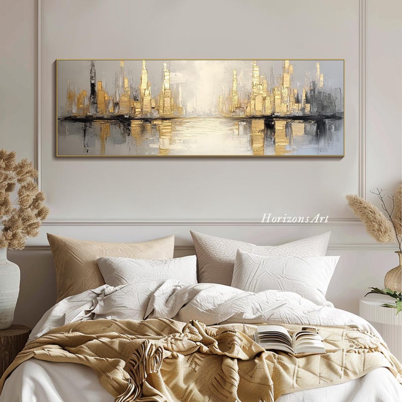 Puede incluir: Pintura abstracta de un paisaje urbano con tonos dorados y grises. La pintura representa un horizonte urbano reflejado en una masa de agua. La pintura est&aacute; enmarcada y colgada en una pared sobre una cama con ropa de cama blanca y una manta de lana marr&oacute;n.