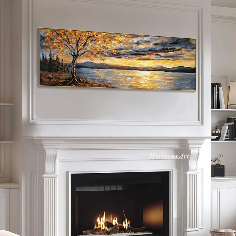 May include: A long, horizontal painting of a lake scene with a tree in the foreground. The tree has yellow leaves and the lake is a deep blue with a golden sunset in the background. The painting is framed in a gold frame and is hanging on a white wall above a fireplace.