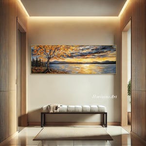 May include: A long, horizontal painting of a lake scene with a tree in the foreground. The tree has yellow and orange leaves. The lake is blue and reflects the sunset sky. The sky is a mix of blue, orange, and purple. The painting is framed in gold.