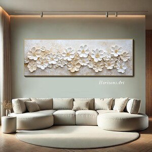 K&ouml;nnte beinhalten: Ein gro&szlig;es, rechteckiges Leinwand-Wandbild mit einem 3D-strukturierten Design aus wei&szlig;en Blumen auf beigefarbenem Hintergrund. Die Blumen sind in einer horizontalen Reihe angeordnet und haben eine erhabene, geformte Optik.