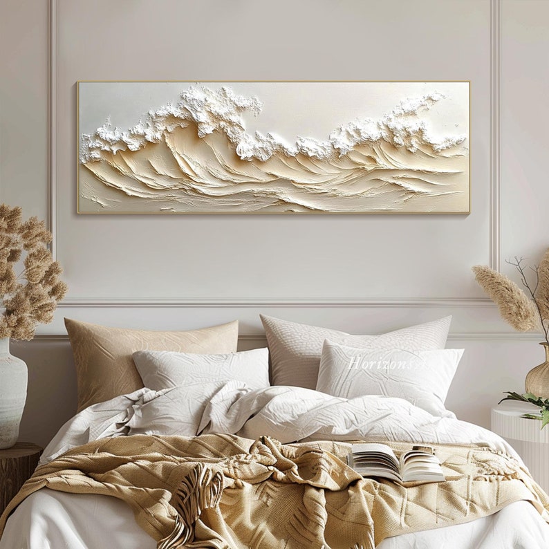 May include: A white and beige textured canvas wall art depicting a wave pattern. The art is framed in a thin gold frame and is hanging on a white wall above a bed with white bedding and a tan throw blanket.