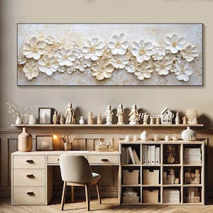 K&ouml;nnte beinhalten: Ein wei&szlig;er und goldener Wandkunstdruck mit floralem Motiv und einem 3D-Textileffekt. Der Druck zeigt eine horizontale Anordnung von stilisierten Blumen mit erhabenen Bl&uuml;tenbl&auml;ttern und einem dezenten goldenen Schimmer. Der Druck ist in einem schwarzen Rahmen gerahmt und wird an einer Wand &uuml;ber einem Schreibtisch mit einem B&uuml;cherregal und dekorativen Figuren ausgestellt.