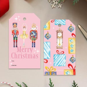 May include: Two pink Christmas gift tags. One features a nutcracker, ballerina, and mouse king with "Merry Christmas" text. The other tag has a clock, presents, and ornaments. Both tags have a hole at the top for string.