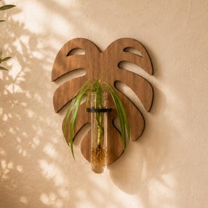 May include: A wall-mounted wooden plant propagation station shaped like a monstera leaf. The brown wood cutout features a central circular opening holding a clear glass test tube with a small green plant inside, secured by a black metal ring.