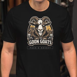 Può includere: T-shirt nera con grafica di testa di capra e la scritta "The Goon Goats Puck'n Hockey". La grafica della capra è nei toni del marrone, beige e bianco. La maglietta è indossata da una persona in palestra.