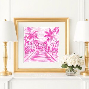 May include: A square framed art print featuring a hot pink ink illustration of a palm tree-lined walkway leading towards a building. The artwork is encased in a thick gold frame and is displayed on a white mantelpiece between two gold lamps.