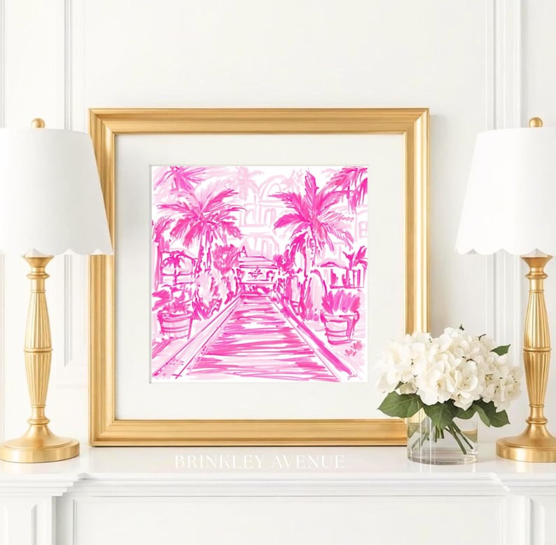 May include: A square framed art print featuring a hot pink ink illustration of a palm tree-lined walkway leading towards a building. The artwork is encased in a thick gold frame and is displayed on a white mantelpiece between two gold lamps.