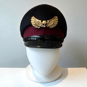 May include: A black and burgundy military-style hat with a gold skull and wing emblem. The hat has a shiny black brim and is displayed on a white mannequin head. The hat's design suggests a gothic or alternative aesthetic.