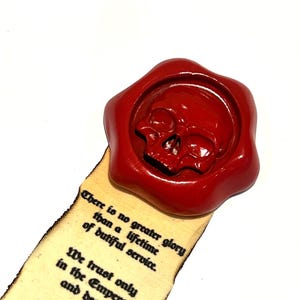 Op de afbeelding: Een rode waszegel met een schedelontwerp is bevestigd aan een houten bladwijzer. De bladwijzer heeft de tekst: "Er is geen grotere glorie dan een leven van plichtsgetrouwe dienst." De bladwijzer is van hout.