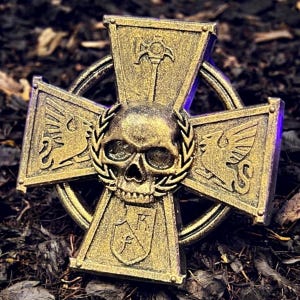 Może przedstawiać: Złoty, ozdobny krzyż z czaszką pośrodku, otoczony wieńcem laurowym. Ramiona krzyża zdobią szczegółowe emblematy i symbole. Projekt ma wygląd postarzanego, antycznego, sugerujący historyczny lub fantastyczny motyw.
