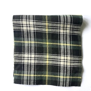 Puede incluir: Tela doblada con un patrón de cuadros clásico. El diseño presenta una base verde oscuro con líneas entrecruzadas en blanco, amarillo y negro. La tela es probablemente lana o un material similar, adecuado para proyectos de ropa o decoración del hogar.