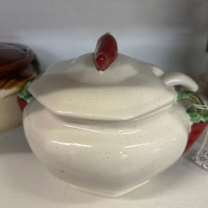 May include: Cream-colored ceramic sugar bowl with a lid and a red chili pepper-shaped handle. The bowl has a rounded shape with red chili pepper accents on the sides. A charming kitchenware item.