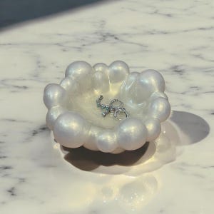 May include: A pearlescent white, bubble-shaped trinket dish. The dish holds two silver hoop earrings and a small, decorative earring. The dish is sitting on a marble surface. Jewelry storage.