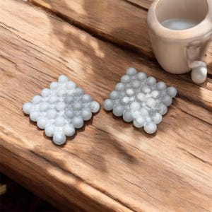 May include: Two square, light gray coasters made of small, round, pearl-like beads. The coasters are arranged on a wooden surface, with a small, off-white ceramic cup in the background. The coasters are designed to protect surfaces from heat and moisture.