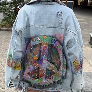 Könnte beinhalten: Eine hellblaue Jeansjacke mit einem Peace-Zeichen, das auf den Rücken gemalt ist. Das Peace-Zeichen ist in verschiedenen Farben bemalt, darunter Rot, Orange, Gelb, Grün, Blau, Lila und Schwarz. Die Jacke hat einen Used-Look mit Rissen und Löchern.