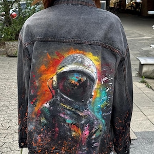 Könnte beinhalten: Dunkelgraue Jeansjacke mit einem lebendigen, farbenfrohen Gemälde eines Astronauten auf dem Rücken. Das Kunstwerk zeigt eine Figur mit Helm vor einem Hintergrund aus orangefarbenen, gelben und blauen Farbspritzern. Die Jacke hat ein klassisches Design mit Kragen und Knöpfen.