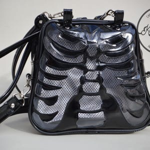 May include: Black faux leather backpack with a clear plastic panel featuring a skeleton ribcage design. The bag has adjustable straps and a top handle. The bag has a logo that says "Kaos Morlina" and "Hecho en Ramos Mejia".