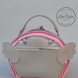 Puede incluir: Un bolso de mano beige brillante con asa superior y un bolsillo horizontal con cremallera. El bolso presenta un borde rosa brillante y dos apliques en forma de alas beige en los laterales. Se observa un logotipo circular con el texto Kaos Morfina.