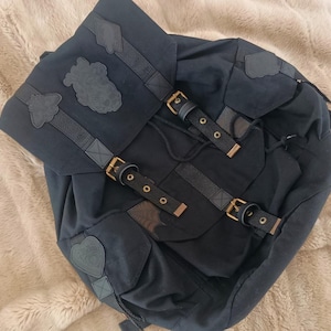 Peut inclure: Un sac à dos bleu marine foncé doté d'un rabat avec un écusson central, deux sangles réglables à boucles et des écussons décoratifs supplémentaires. Le sac comprend une fermeture à cordon et des détails en métal doré.