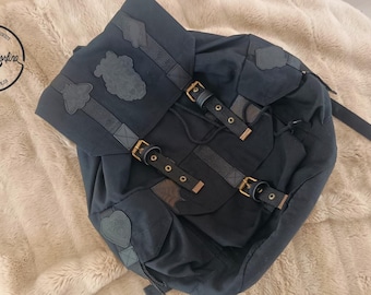 May include: A dark navy blue backpack featuring a fold-over flap with a central patch, two adjustable buckled straps, and additional decorative patches. The bag includes a drawstring closure and gold-toned metal hardware accents.