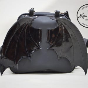 May include: Black backpack with glossy, bat-wing shaped accents. The bag has a rounded shape and features adjustable straps and a top handle. The wings are a shiny black, contrasting with the matte finish of the bag. The bag has the text "Carteras y Accesorios Kaos Morfina Desde 2007 Hecho en Ramos Mejia".