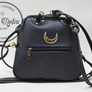 May include: A black faux leather backpack with a gold crescent moon design. The bag has a single adjustable strap and a zippered pocket on the front. The bag has the text "Kaos Morfina" and "Hecho en Ramos Arizpe" on the front.