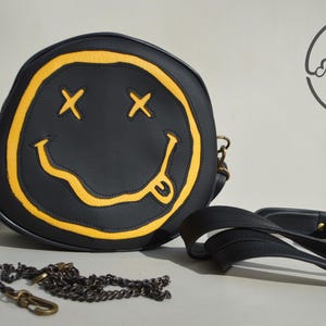 May include: A black round crossbody bag with a yellow smiley face design. The bag has a long adjustable strap and a chain strap. The bag is made of leather and has a zipper closure.