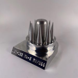 May include: A metallic silver object with a square base and a cylindrical top. The top features a series of conical spikes surrounding a central, rounded cylinder. The base has engraved text: "UDEN-SEE Mk-3 RV/W33".