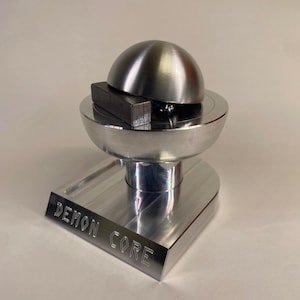 May include: A polished metal sculpture with a semi-spherical top and a rectangular base. The base is engraved with the words "DEMON CORE". A rectangular metal piece rests on the sculpture's bowl-shaped center.