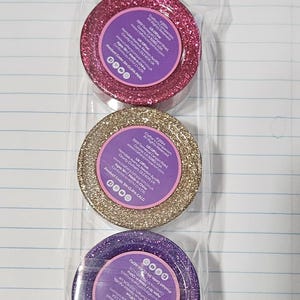 May include: Three rolls of glitter tape in pink, gold, and purple, each packaged in clear plastic. The tape rolls have a circular label with text. The product code is SN-GLIPA-PEBL, SN-GLIPA-CALG, and SN-CORA REPU.