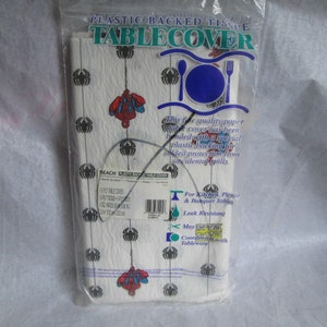 May include: A package of white plastic-backed paper table cover. The table cover has a pattern of black spiders and red and blue Spider-Man figures. The packaging includes text that reads "Plastic Backed Tissue Tablecover".