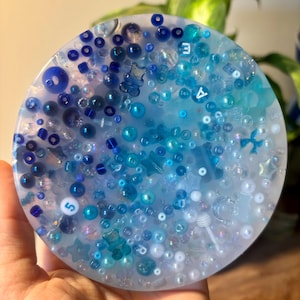 May include: A round, clear resin coaster with a variety of blue, white, and iridescent beads and charms embedded in it. The coaster has a light blue, white, and dark blue gradient.