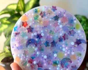 May include: A round, clear resin coaster with a whimsical design featuring iridescent stars, moons, and beads in shades of purple, pink, blue, and yellow.