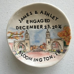 Op de afbeelding: Een witte keramische ringenschotel met een handgeschilderde illustratie van een gebouw met twee torens. De schotel is beschreven met de tekst "James & Ashley Engaged December 23, 2016 Bloomington, IN."