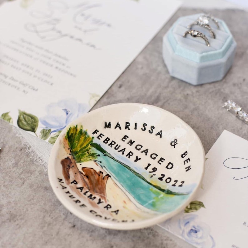 Op de afbeelding: Een witte keramische ringenschotel met een geschilderd tafereel van een strand met palmbomen en de woorden "Marissa & Ben Verloofd Februari 19, 2022" en "Palmyra Nature Cove" erop geschreven.