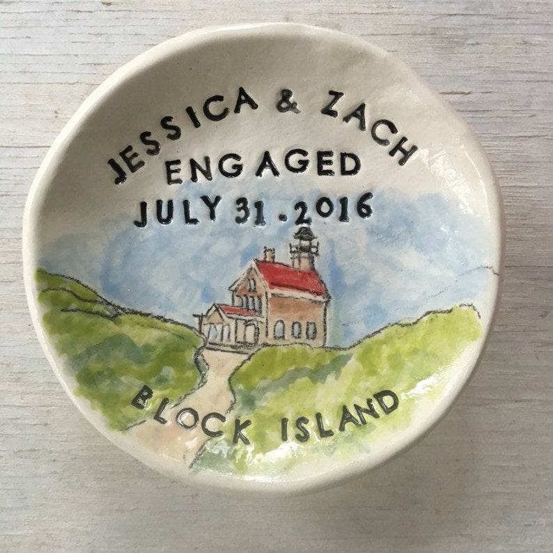 Op de afbeelding: Een witte keramische ringenschotel met een handgeschilderd ontwerp van een vuurtoren op Block Island. De schotel is beschreven met de tekst "Jessica & Zach Verloofd op 31 juli 2016".