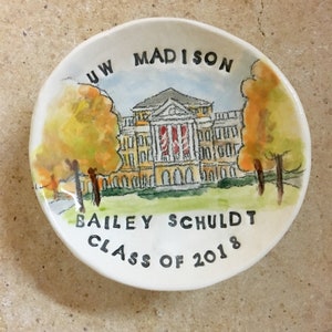 May include: A white ceramic dish with a hand-painted image of a building with the text "UW MADISON" above it. The building is yellow with red accents and is surrounded by trees. The text "BAILEY SCHULDT CLASS OF 2018" is written below the building.