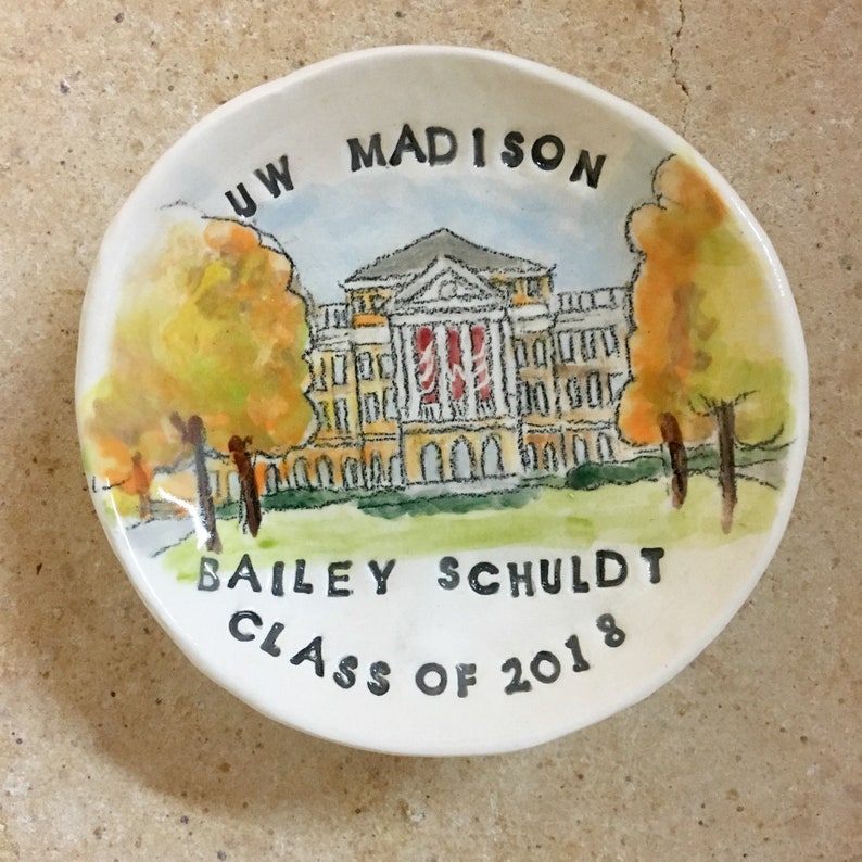 May include: A white ceramic dish with a hand-painted image of a building with the text "UW MADISON" above it. The building is yellow with red accents and is surrounded by trees. The text "BAILEY SCHULDT CLASS OF 2018" is written below the building.