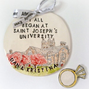 May include: A white ceramic ornament with a hand-painted illustration of a university building and the text "It all began at Saint Joseph's University". The ornament also includes the names "Dan & Kristina" and a small ceramic ring with a diamond.