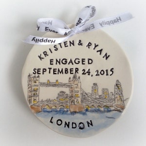 K&ouml;nnte beinhalten: Wei&szlig;er Keramik-Anh&auml;nger mit einer Schwarzwei&szlig;-Illustration der Tower Bridge in London. Der Anh&auml;nger ist mit dem Text "Kristen & Ryan Verlobt am 24. September 2015 London" verziert.