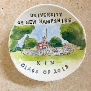 May include: A white ceramic plate with a watercolor painting of a red brick building with a green roof and a white steeple. The building is surrounded by green trees and a stone wall with the words "University of New Hampshire" on it. The plate also has the words "Class of 2018" and "Kim" written on it.