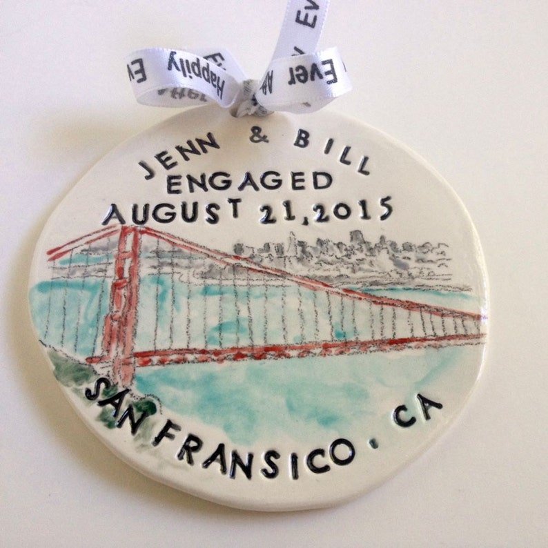K&ouml;nnte beinhalten: Ein wei&szlig;er Keramik-Anh&auml;nger mit einem handgemalten Bild der Golden Gate Bridge in San Francisco. Der Anh&auml;nger ist mit dem Text "Jenn & Bill Verlobt am 21. August 2015 San Francisco, CA" verziert.