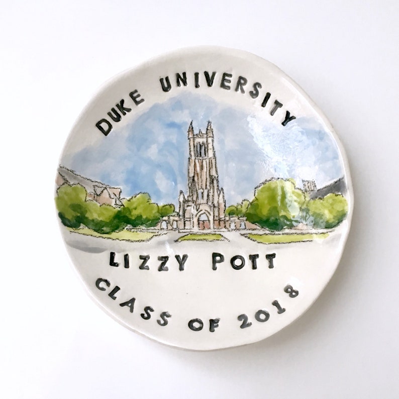 May include: A white ceramic dish with a watercolor painting of a building and trees. The text "DUKE UNIVERSITY" is written around the top edge of the dish. The text "Lizzy Pott CLASS OF 2018" is written on the bottom of the dish.