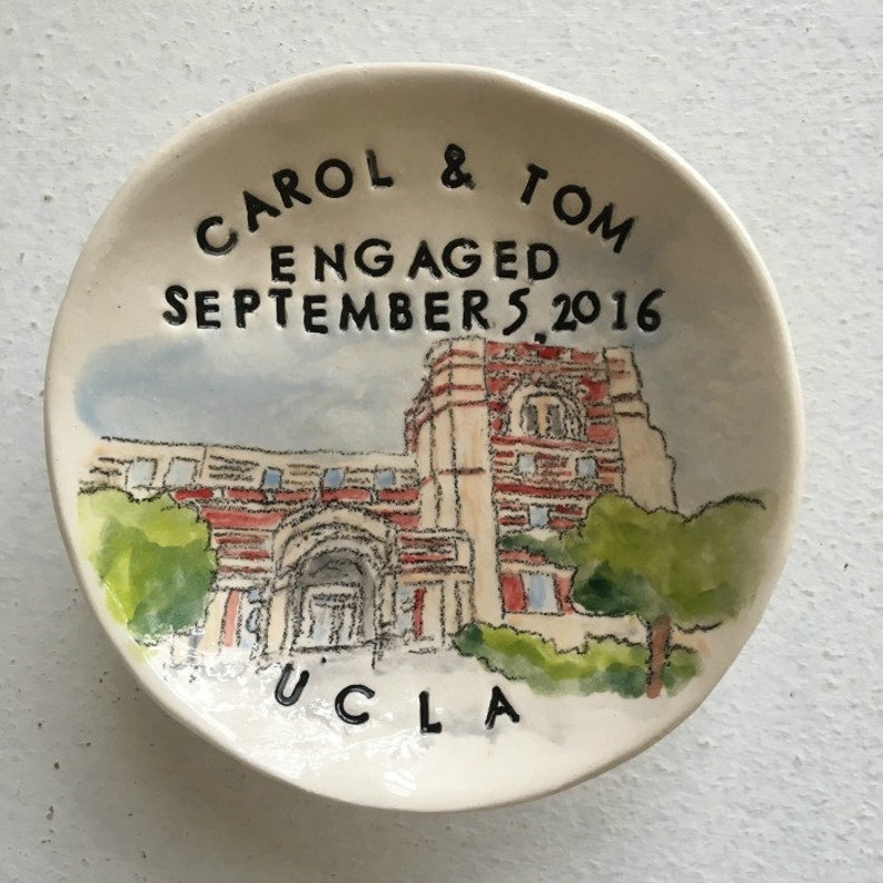 Op de afbeelding: Een witte keramische schaal met een aquarel van een gebouw en de tekst "CAROL & TOM ENGAGED SEPTEMBER 5, 2016 UCLA".
