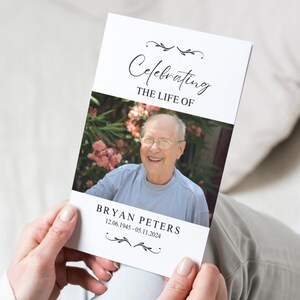 Op de afbeelding: Een wit gedenkboekje met een zwarte rand en een bloemmotief. Het boekje bevat een foto van een lachende man met een bril en de tekst "Celebrating the Life of Bryan Peters 12.06.1945 - 05.11.2024".