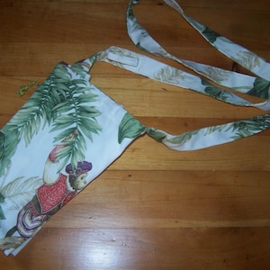 May include: A white fabric bag with a green and brown tropical print featuring a monkey wearing a red and white outfit. The bag has a long white strap.