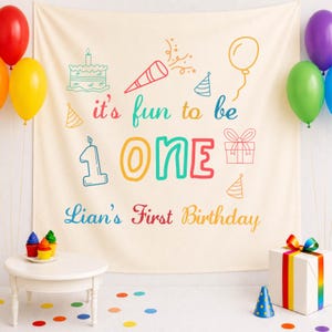 Puede incluir: Una pancarta de cumpleaños color crema con texto e ilustraciones coloridas. La pancarta dice "it's fun to be ONE" y "Lian's First Birthday". También se ven globos y una caja de regalo.