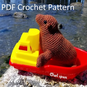 May include: A brown crocheted sea lion sitting in a red and yellow toy boat. The boat is on a rock covered in barnacles. The text "Owl spun" is visible on the boat.