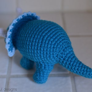 May include: A crocheted blue triceratops dinosaur toy with white accents. The toy has a frill around its head and a long tail.